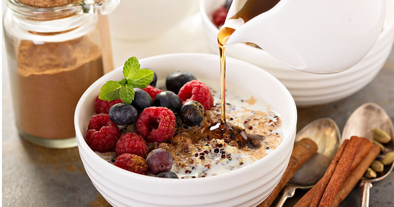 A bowl of cereal with fruit on top. Honey is being drizzled into the wol. A container of cinnamon is in the background.