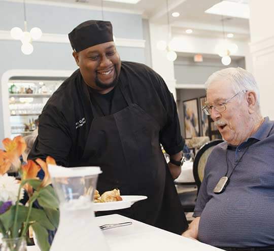 Chef at Heartis location presents a resident with a special meal while the resident looks on appreciatively.