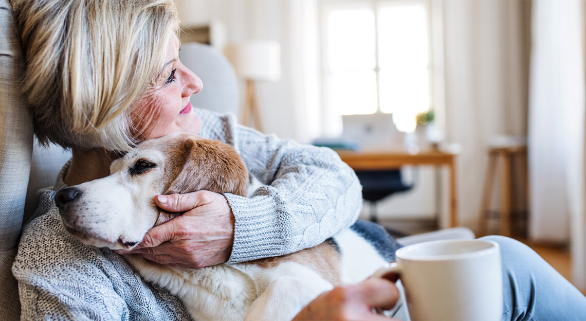 A senior women sitting in a chair, wearing a gray sweater, hugging her Labrador dog while holding a white mug and smiling