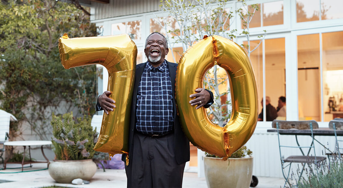 A senior man holding large gold number 7 and 0 giant balloons, standing in patio area and laughing out loud